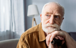 Senior man looking off into distance sitting on couch with hands and head resting on walking stick, forgetful, memory loss