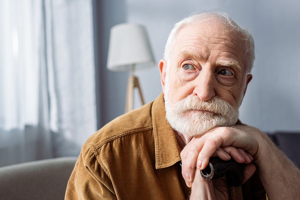 Senior man looking off into distance sitting on couch with hands and head resting on walking stick, forgetful, memory loss