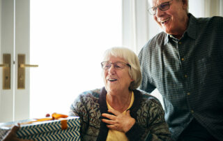Senior woman receiving gift, husband standing behind her near window